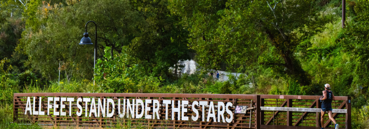 Runner in dark blue with white hat running across All Feet Stand Under the Stars greenway bridge in Asheville.